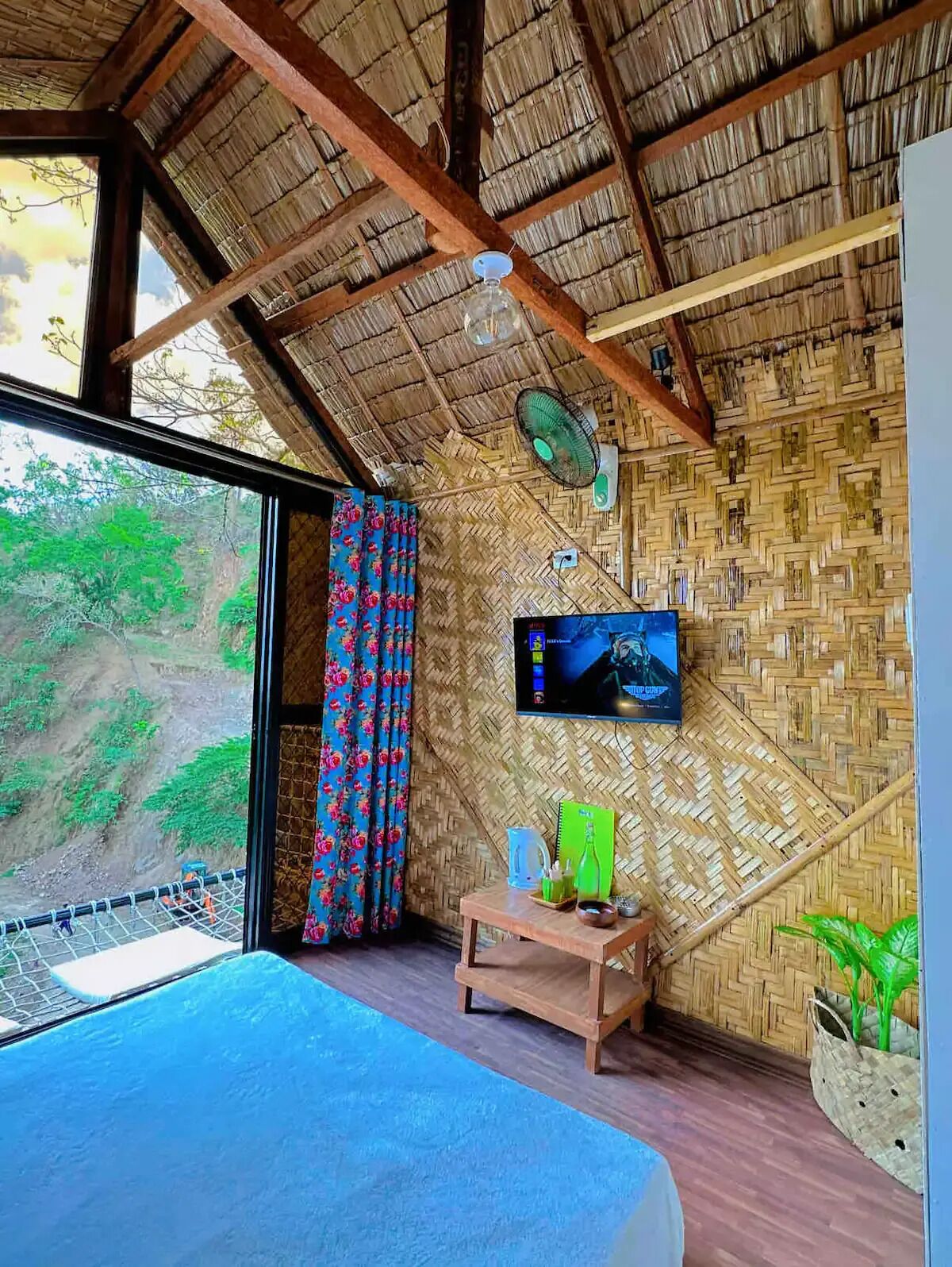 Una habitación con techo de paja, una cama, un televisor, un ventilador y vistas a la vegetación.