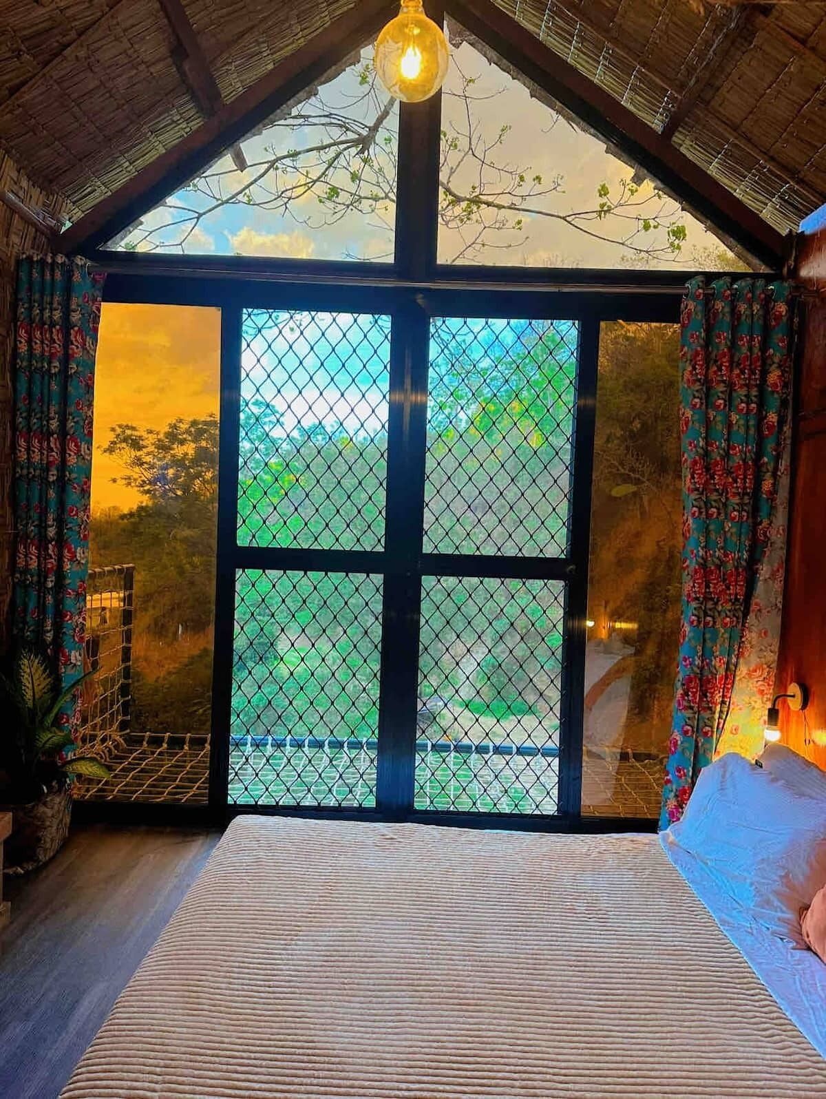 Een kamer met een schuine kap, een bed, een hanglamp en een groot raam met hor.