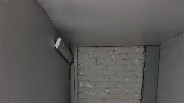 A small, narrow room with a single bed, a brick wall, and a white air conditioning unit mounted on the wall.