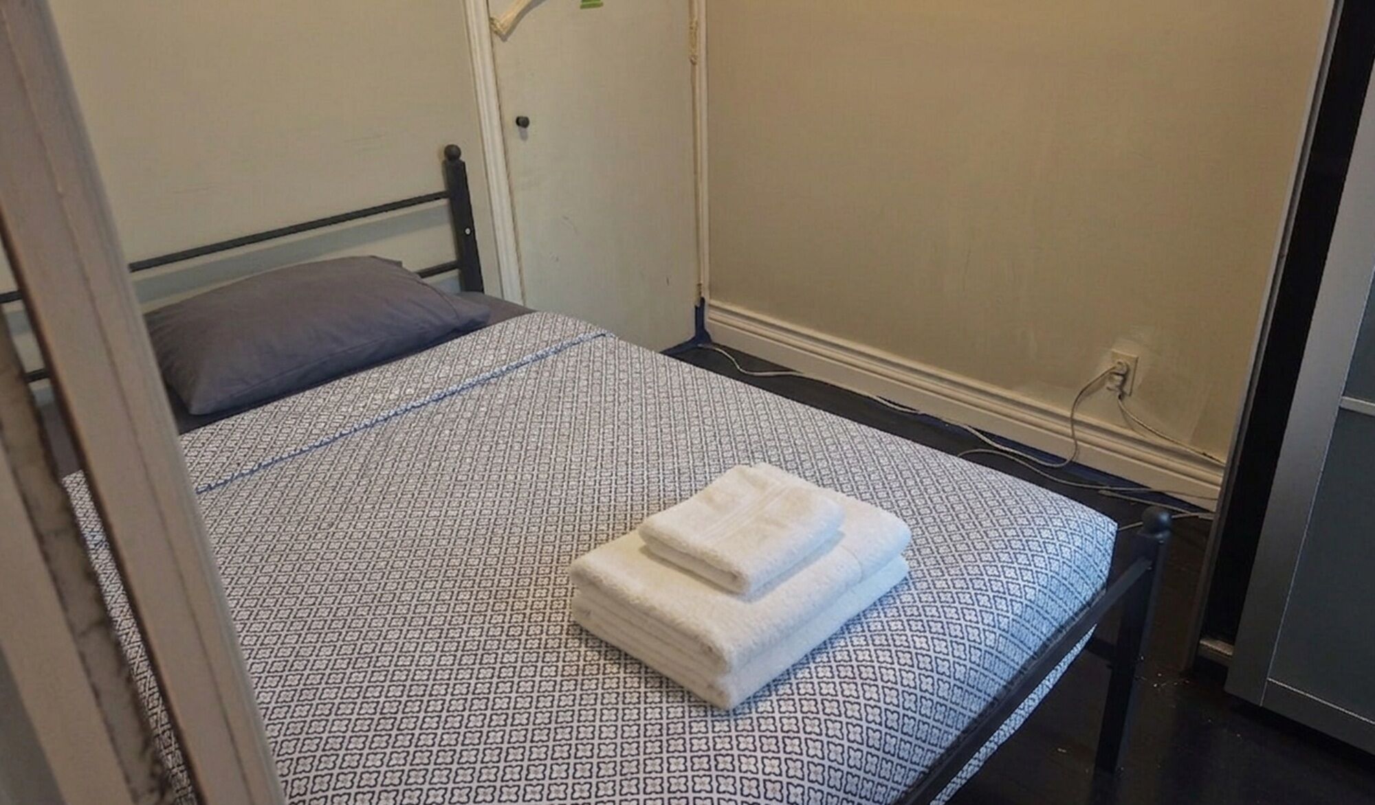 Habitación pequeña con una cama individual, una colcha estampada, una toalla blanca doblada y una puerta a la izquierda.