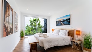 A modern bedroom with a large bed, bedside tables, a bench, and a view of greenery through a sliding door. - T3 Casa Bonita Vacation Apartment in Nazaré (Sítio da Nazaré)
