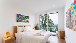 A modern bedroom with a large bed, a cityscape painting, and a view through a sliding glass door. - T3 Casa Bonita Vacation Apartment in Nazaré (Sítio da Nazaré)