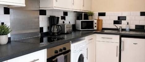 Cocina compacta con armarios blancos, encimera negra, microondas, cafetera, lavadora y refrigerador.