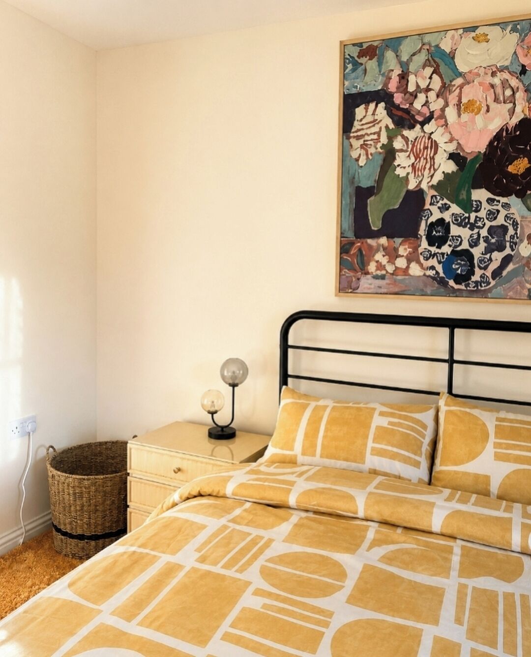 A bedroom with a bed, a nightstand, a lamp, a wicker basket, and a painting on the wall.
