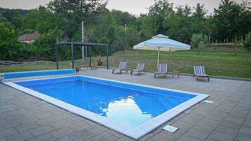 A rectangular swimming pool with a tiled deck, surrounded by lounge chairs and a covered seating area.