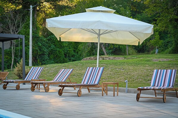 A patio with lounge chairs and a large umbrella, surrounded by greenery.