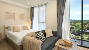 Quarto de hotel com cama, sofá, mesa com livros e varanda com vista.