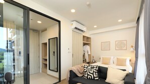 Quarto de hotel moderno com porta de vidro que dá para uma varanda, sofá preto com almofadas estampadas, mesa de cabeceira com livros, cama com roupa de cama branca e ar condicionado fixado na parede.