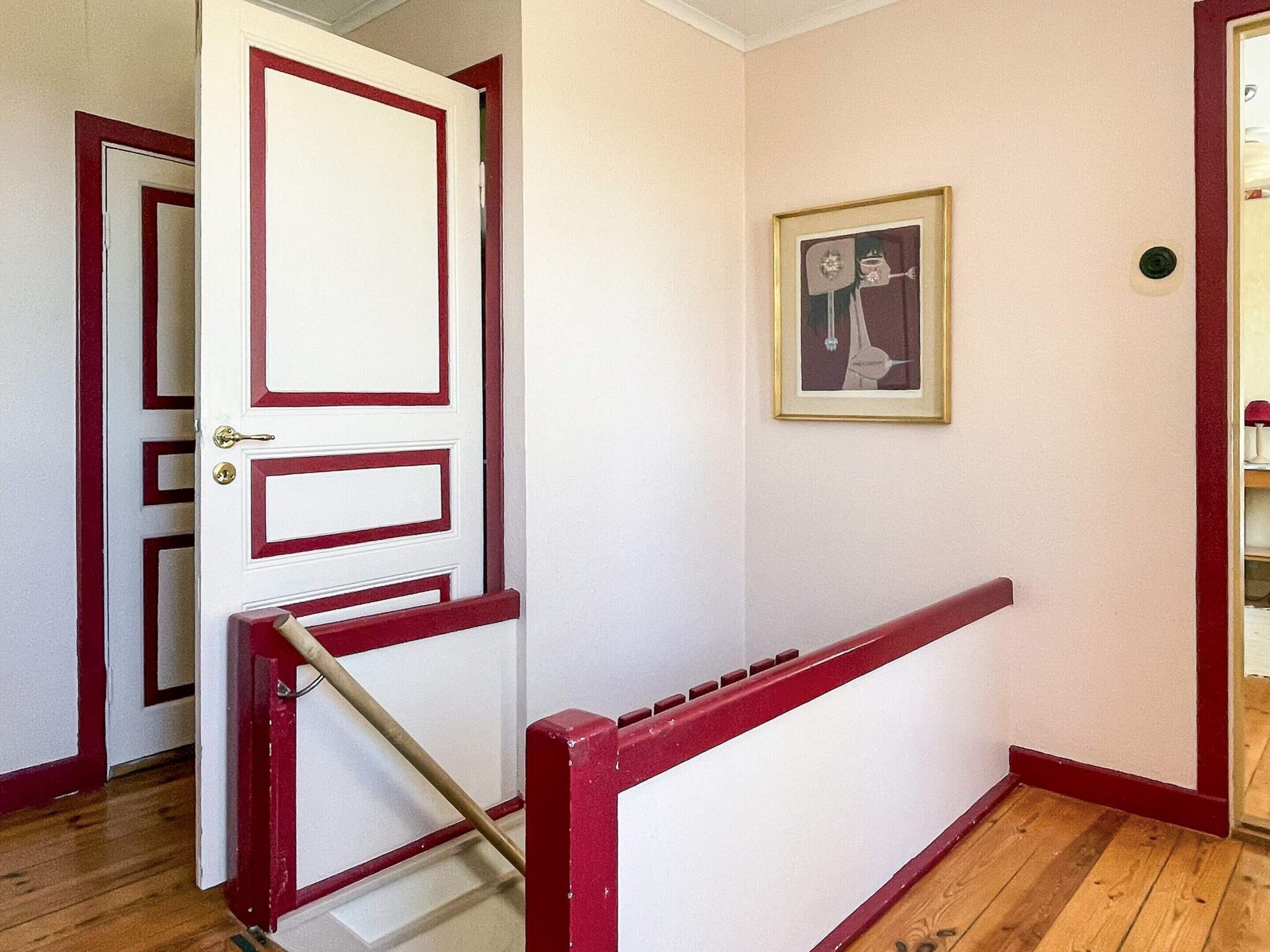 A hallway with a white door with red trim, a wooden floor, and a framed picture on the wall.