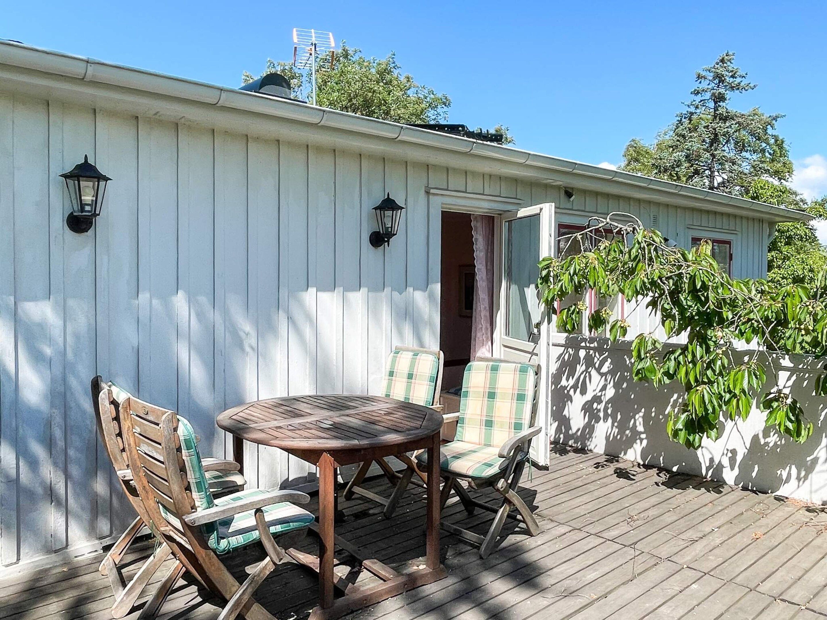 Une terrasse en bois avec une table ronde et des chaises, un bâtiment blanc avec une porte, et un arbre aux feuilles vertes.