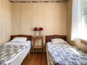 Ein Zimmer mit zwei Betten, jeweils mit einem Holzkopfstück, einem kleinen Nachttisch mit zwei Lampen und einem Fenster mit durchscheinenden Vorhängen.