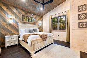 A bedroom with a large bed, wooden walls, a ceiling fan, and a window with curtains. - NEW! Pickleball, Hot Tub, ATV Trails, Outdoor Movies & Mini Golf (Broken Bow)
