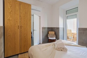 A bedroom with a wooden wardrobe, a bed with white linens, a wicker chair, and a view of the outdoors. - Setúbal Downtown Fonte Nova, By Home Match (Setúbal)