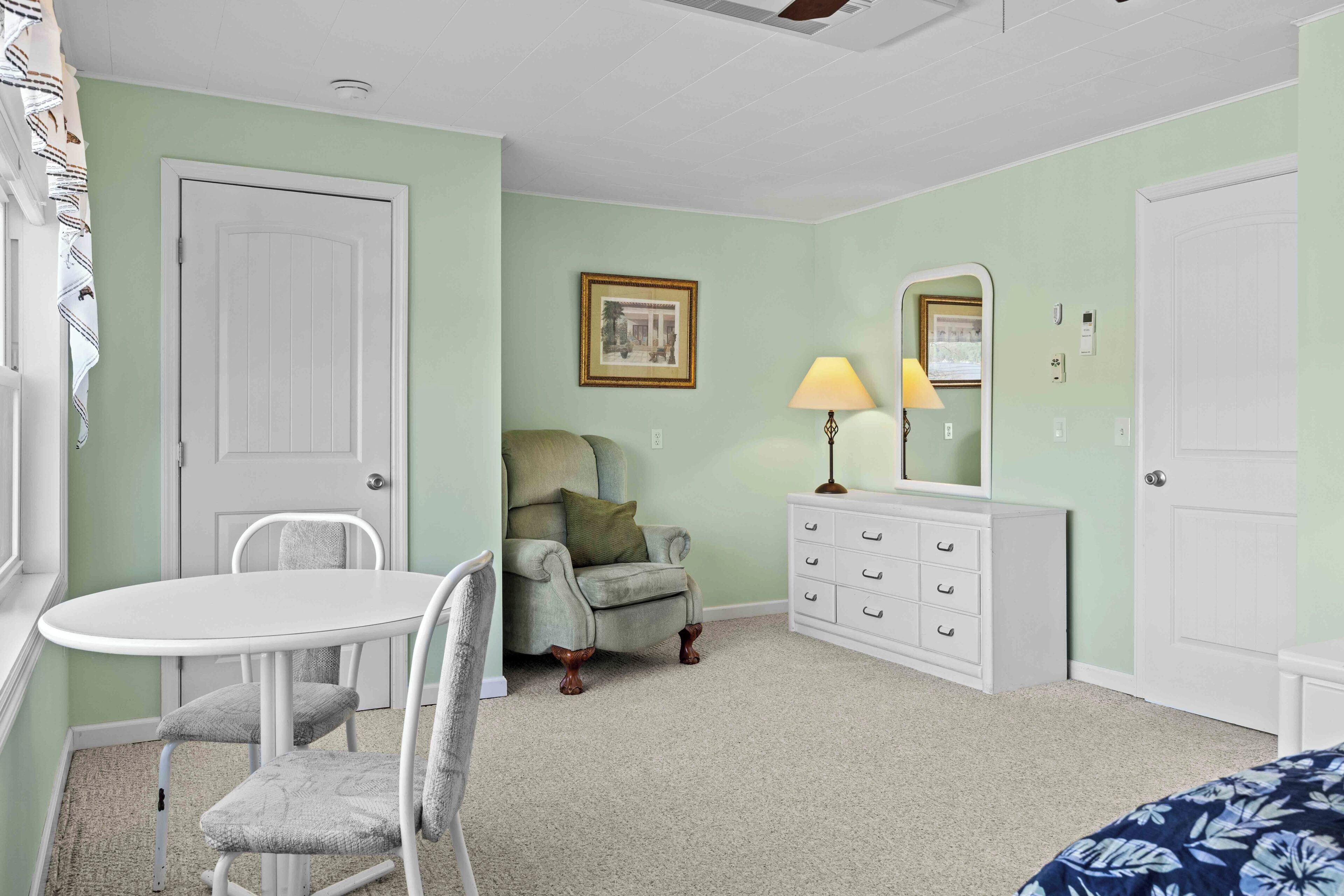 Una habitación con una puerta blanca, un sillón verde, una mesita con sillas, un aparador y una lámpara.