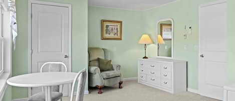 Una habitación con una puerta blanca, un sillón verde, una mesita con sillas, un aparador y una lámpara.