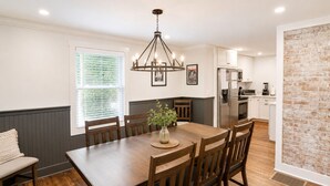A dining area with a wooden table, chairs, a chandelier, and a kitchen in the background.