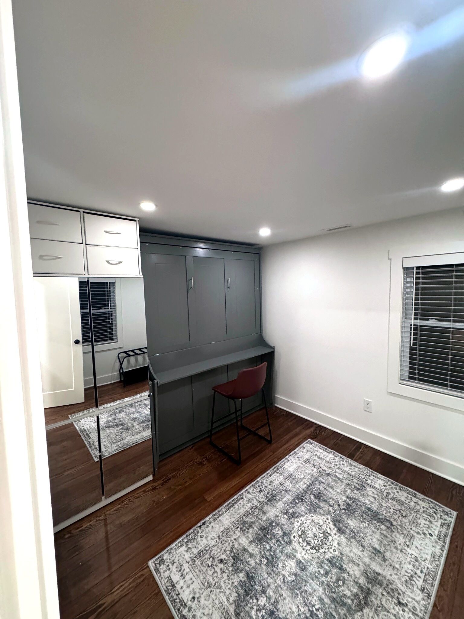 A compact room with a kitchenette, a desk, a chair, and a large rug.