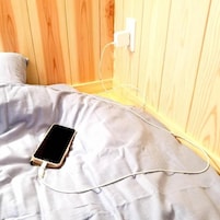 Una cama con un teléfono inteligente cargando, un enchufe blanco en la pared y un cabecero de madera.