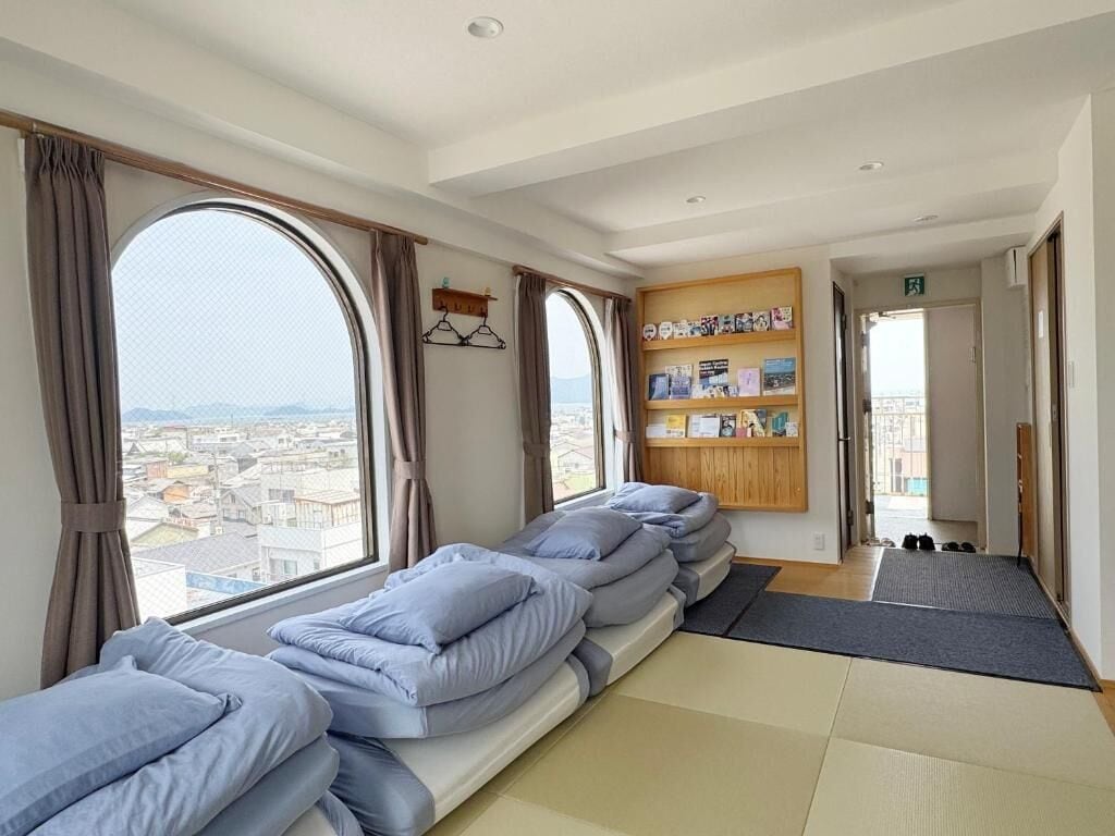 A hotel room with a large window, a sofa bed, a bookshelf, and a shoe rack.