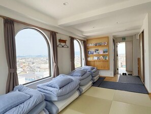 A hotel room with a large window, a sofa bed, a bookshelf, and a shoe rack.