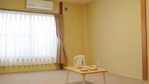 A room with a beige carpet, a small wooden table with a kettle and cups, a window with sheer curtains, and an air conditioning unit on the wall. - Shimanami Guest House Orangenokaze - Hostel  (Imabari)