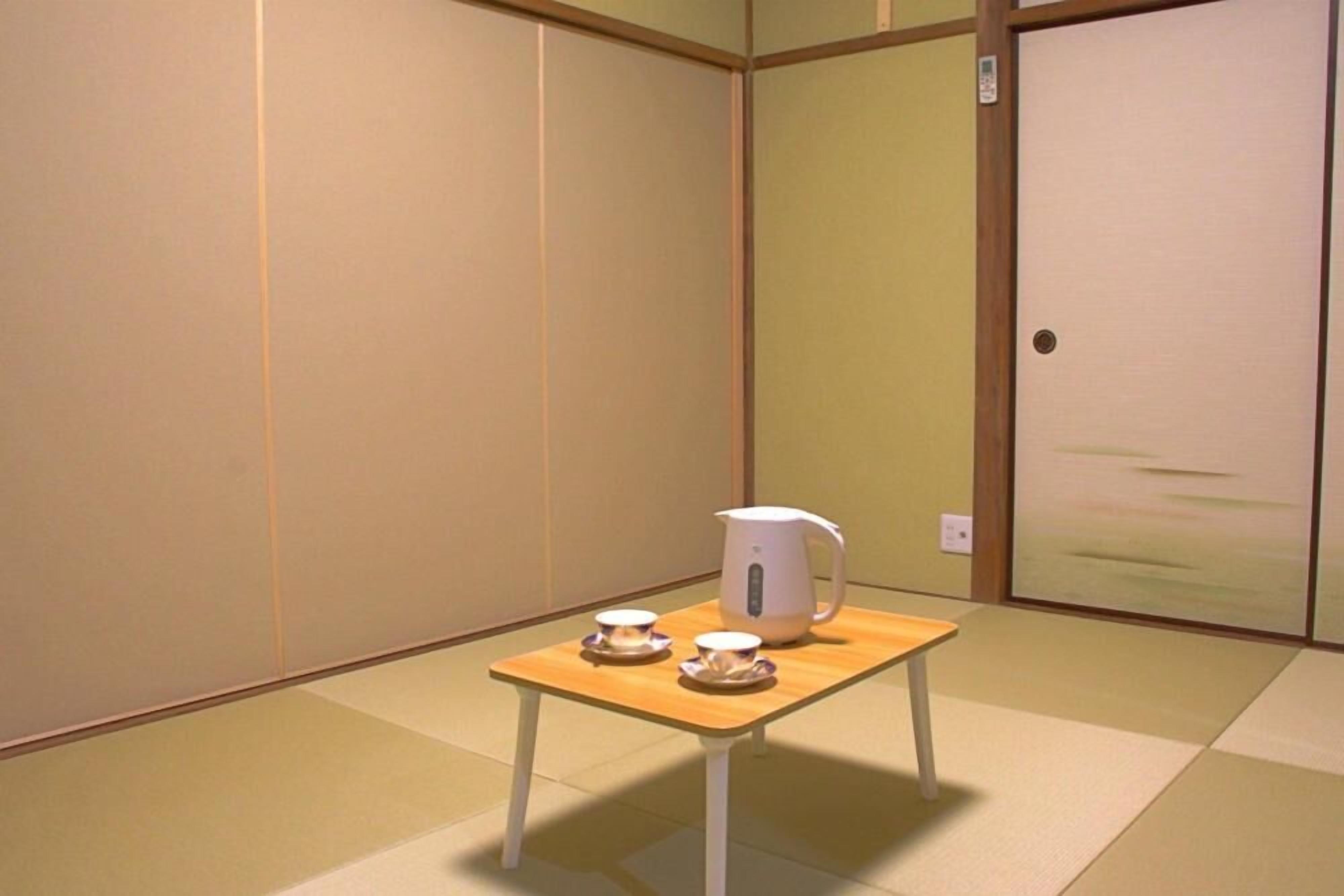 Une pièce avec un sol recouvert de tatamis, une petite table avec une bouilloire et deux tasses, et une porte coulissante.