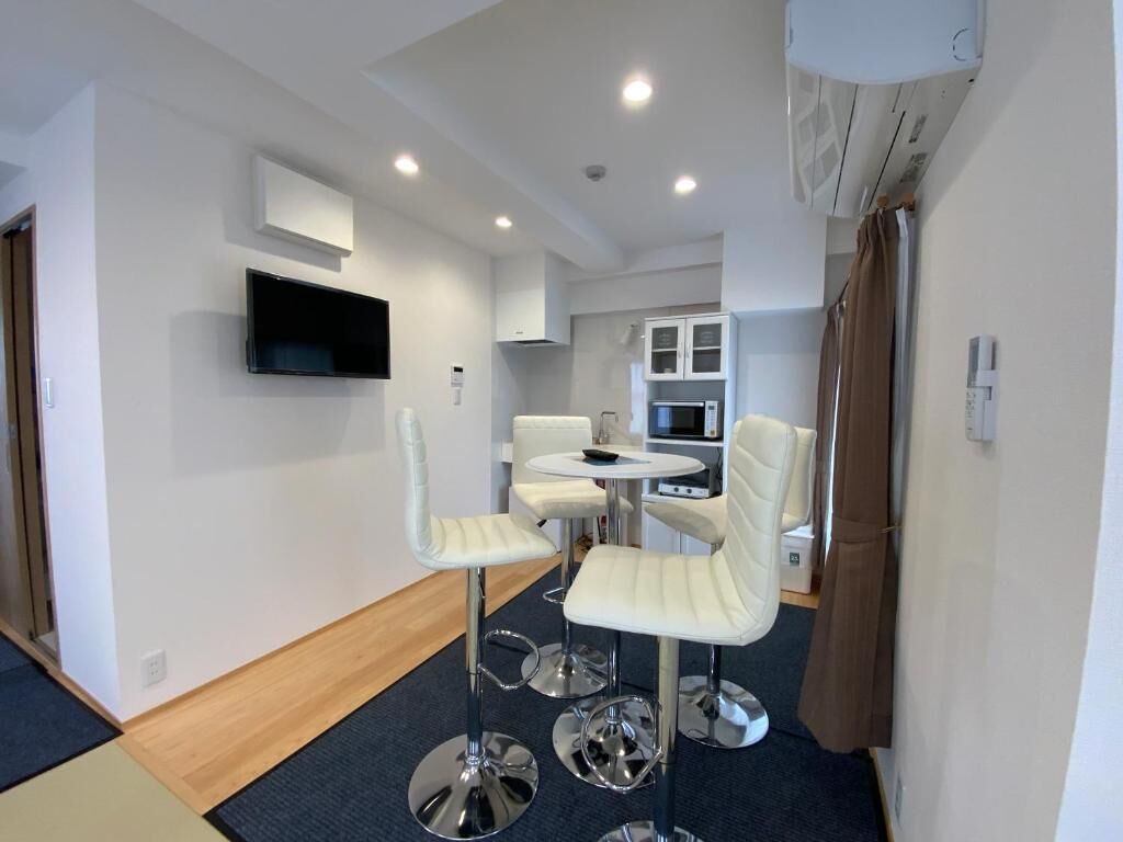 Una habitación de hotel moderna con televisión de pantalla plana, una zona de comedor con sillas blancas, una mesita redonda y una kitchenette con armarios y microondas.