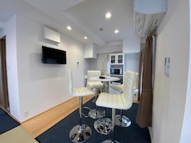 Una habitación de hotel moderna con televisión de pantalla plana, una zona de comedor con sillas blancas, una mesita redonda y una kitchenette con armarios y microondas.