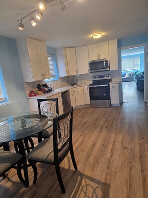 A modern kitchen with white cabinets, a microwave, and a dishwasher. There is a dining table with chairs in the foreground. - Casa Azul : Cozy Retreat Near Obama Library (Chicago)