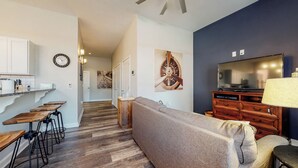 A modern living room with a sofa, wooden coffee table, and bar stools. A TV and wooden cabinet are in the background. - Cozy 1-bedroom condo in downtown Louisville with free parking and gym access (Louisville)