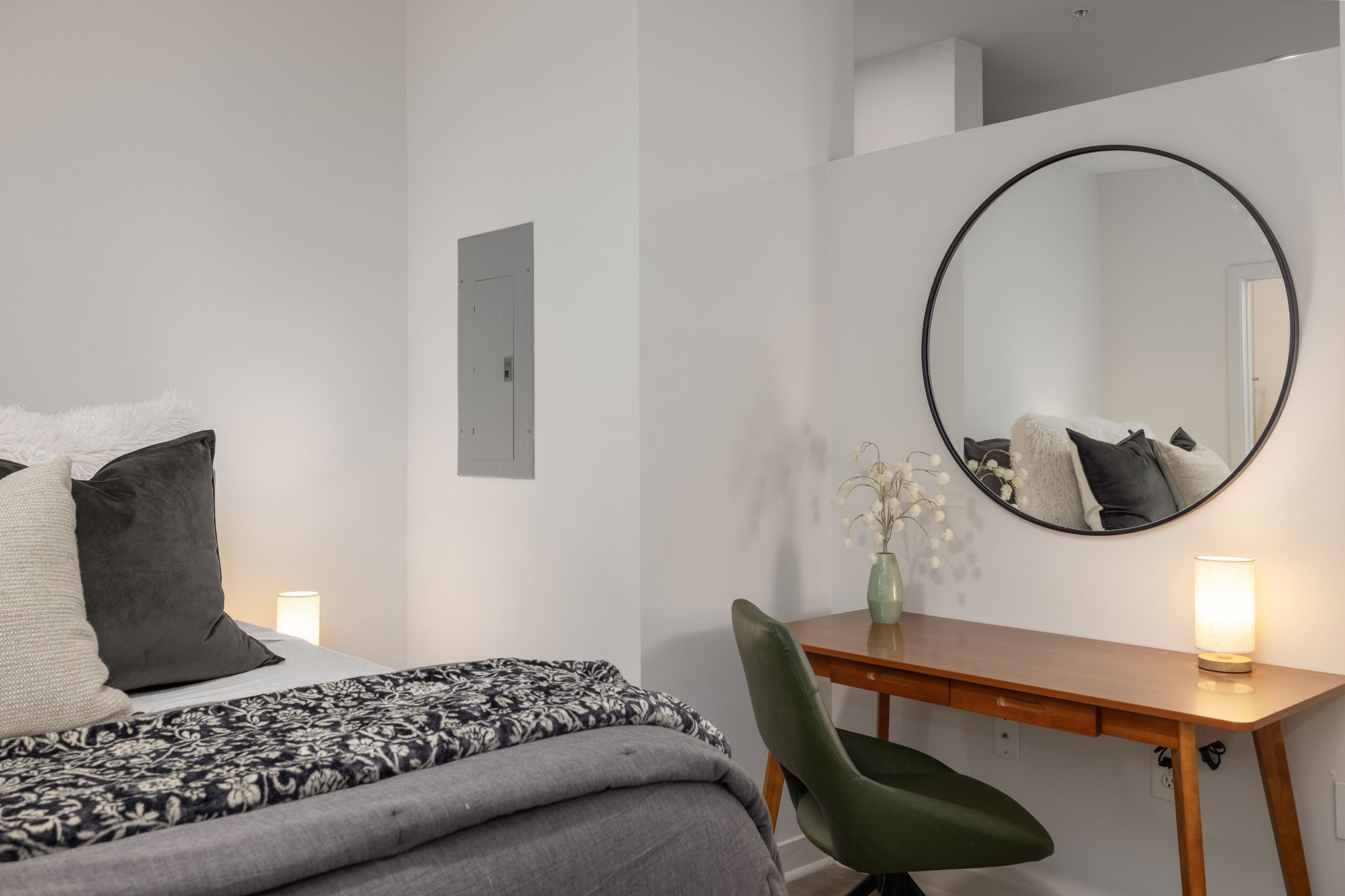 Une chambre à coucher avec un lit, un bureau, une chaise, un miroir rond et un vase avec des fleurs.