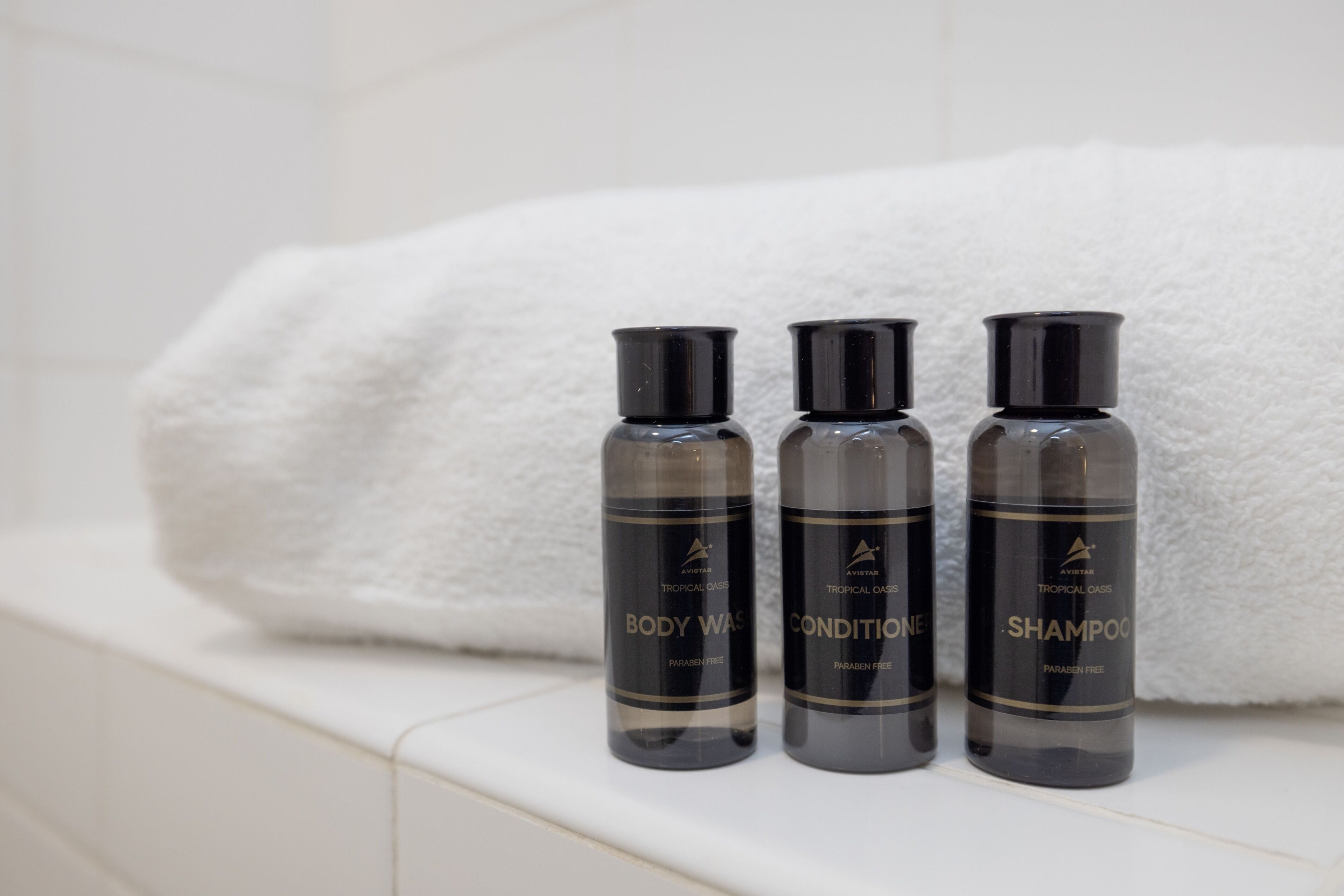 Drei Flaschen Duschgel, Conditioner und Shampoo auf einer weißen Fliesenfläche, im Hintergrund ein weißes Handtuch.