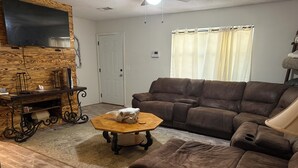 A living room with a brown leather sofa, a wooden coffee table, a flat-screen TV mounted on a wooden wall, and a door.