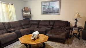 A living room with a brown leather sectional sofa, a wooden coffee table with a skull, a small table with a lamp, and a framed picture on the wall.