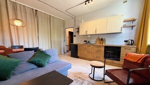 Ein modernes Wohnzimmer mit einer Couch, einem Tisch, einem Sessel und einer Küchenzeile mit Schränken und Geräten.
