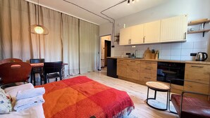 Ein modernes Zimmer mit einem Bett, einem Esstisch, Stühlen, einer Küchenzeile mit Schränken und Geräten sowie einem großen Fenster mit Vorhängen.