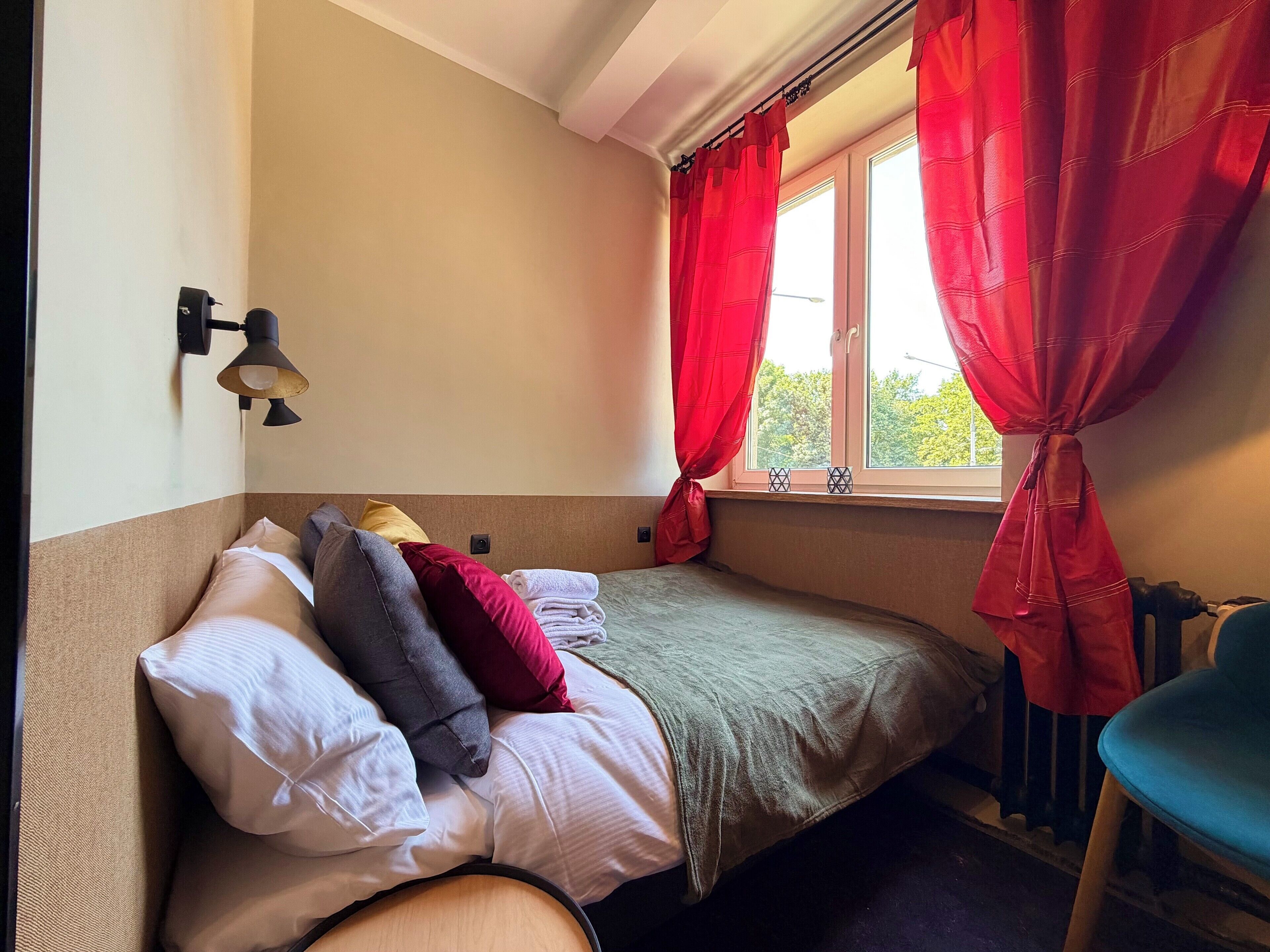 Un dormitorio con cama, almohadas, una ventana con cortinas rojas, un radiador y una lámpara de pared.