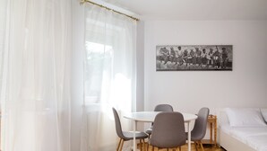 Une pièce avec un lit blanc, une table de salle à manger ronde entourée de quatre chaises, un rideau blanc et une photographie en noir et blanc accrochée au mur.