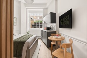 Een compacte kamer met een bed, een kleine kitchenette, een eethoek met een tafel en stoelen, en een raam met uitzicht op gebouwen.