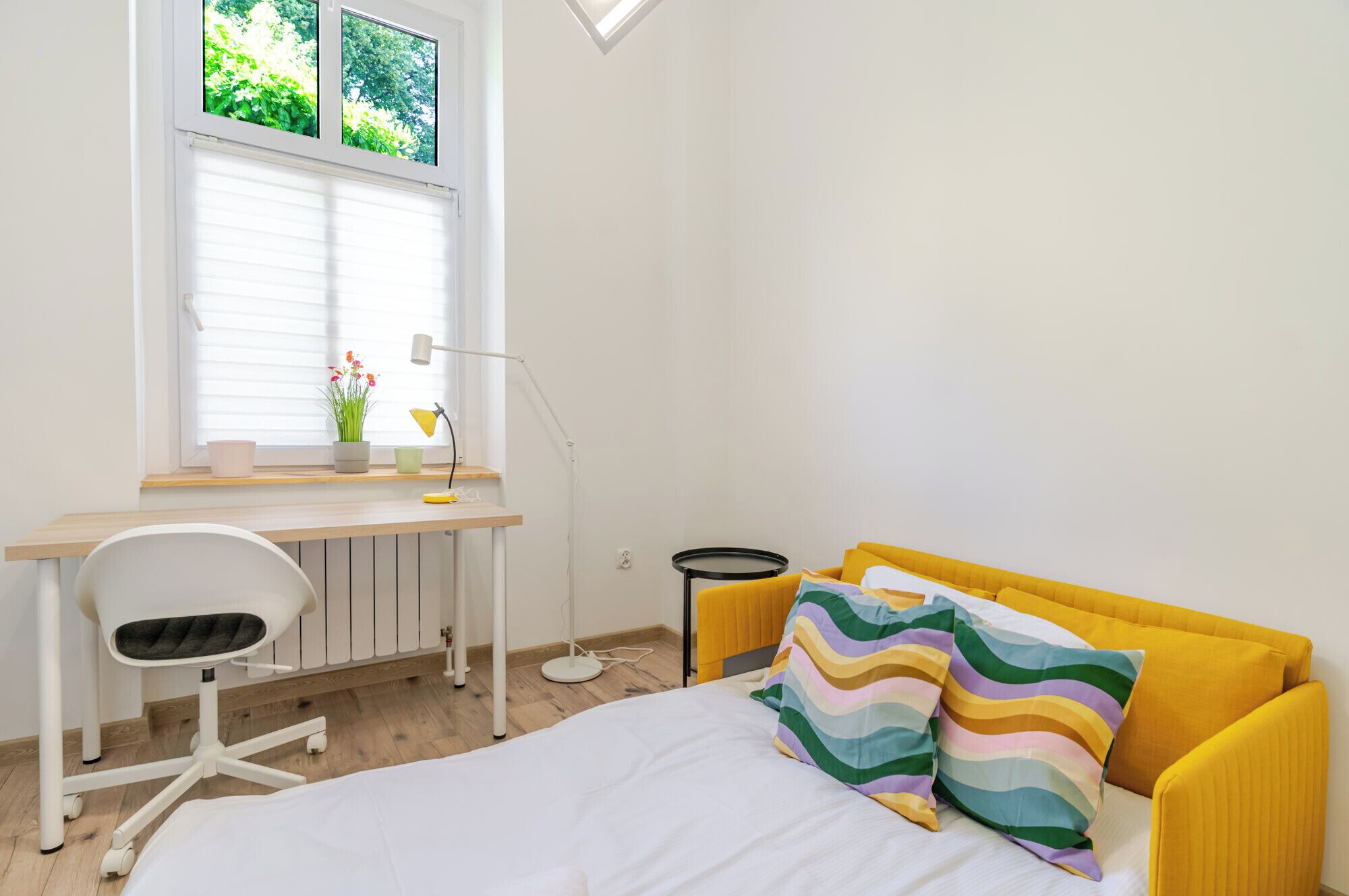 Een slaapkamer met een bed, een bureau, een stoel, een raam met jaloezieën en een kleine tafel.