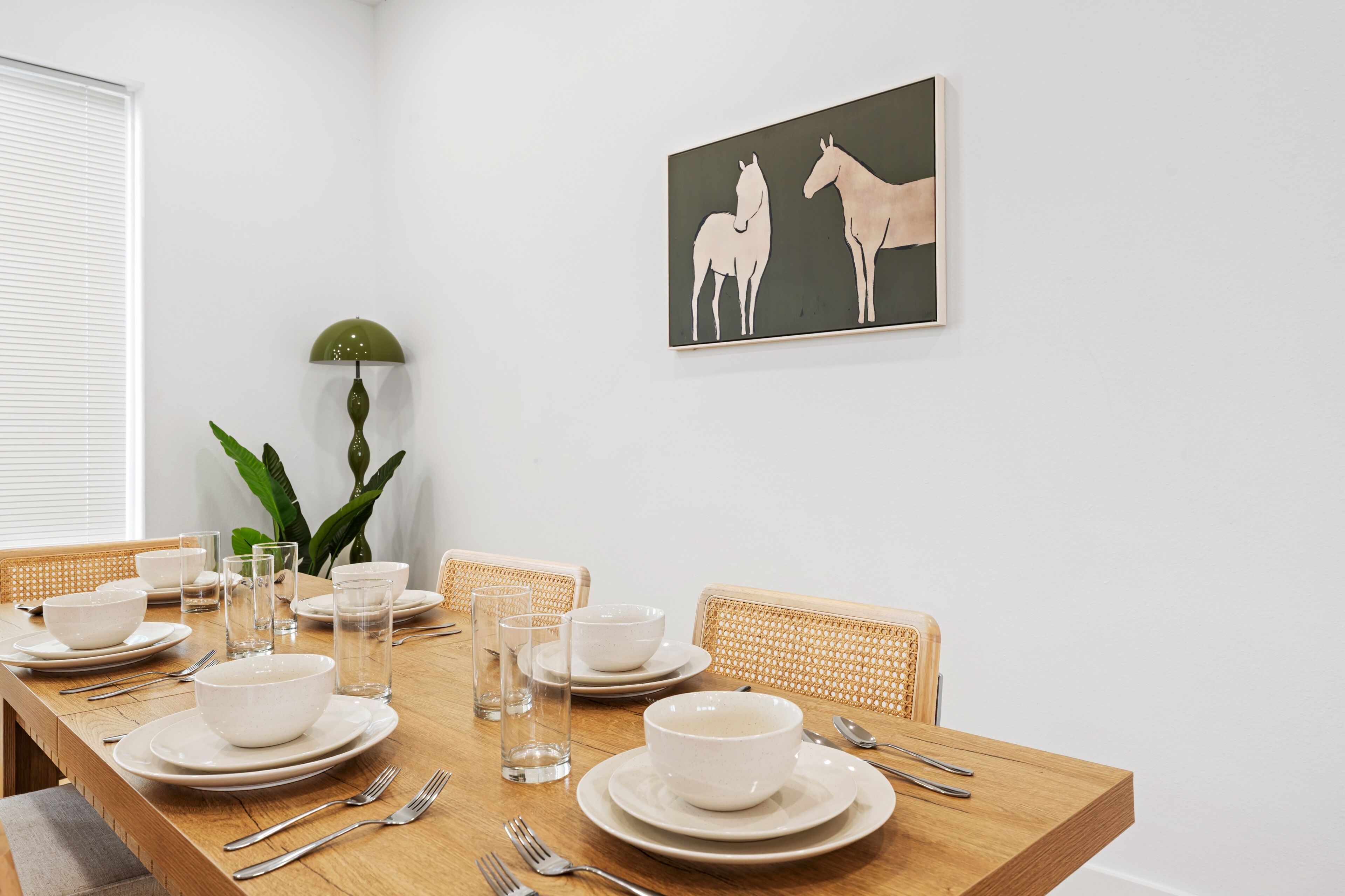 A dining area with a wooden table set for four, white plates, bowls, and cutlery, a green floor lamp, and a wall-mounted painting of horses.