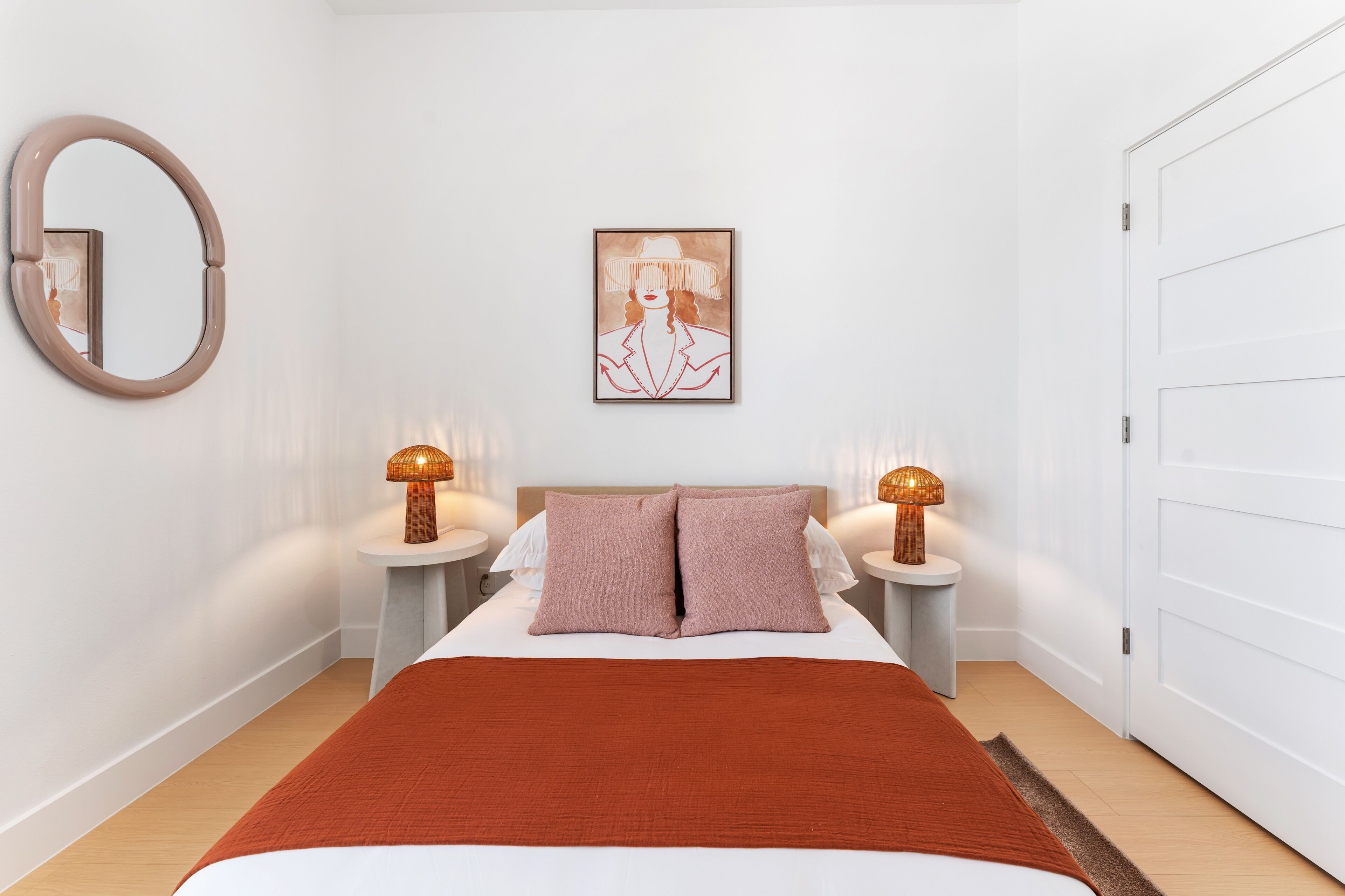 Un dormitorio con una cama, dos mesitas de noche, un espejo montado en la pared, una obra de arte enmarcada y una puerta blanca.