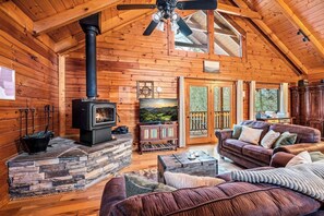 Ein Interieur einer Blockhütte mit Kamin aus Stein, einem Ledersofa, einem Fernseher und einer reizvollen Aussicht durch die Glastüren.