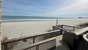 Blick auf einen Strand von einer Holzterrasse aus, mit einem Pier in der Ferne.