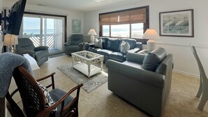 Ein Wohnzimmer mit Sofa, Sesseln, einem Couchtisch und einem Fernseher. Ein Fenster bietet Blick auf den Strand.
