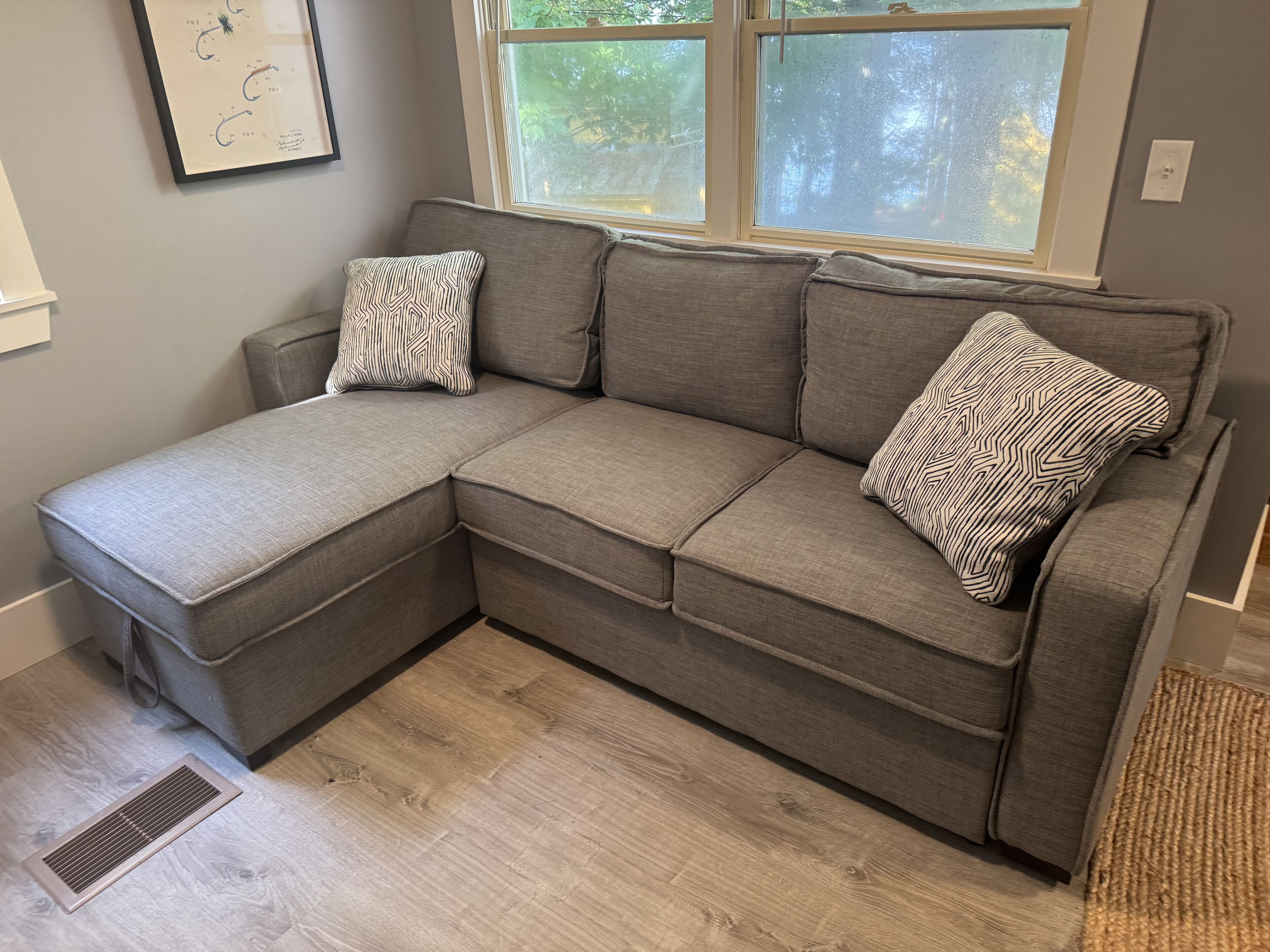 Ein graues Ecksofa mit gemusterten Kissen, ein Fenster mit Blick auf Bäume und ein heller Holzboden.