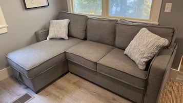 Ein graues Ecksofa mit gemusterten Kissen, ein Fenster mit Blick auf Bäume und ein heller Holzboden.
