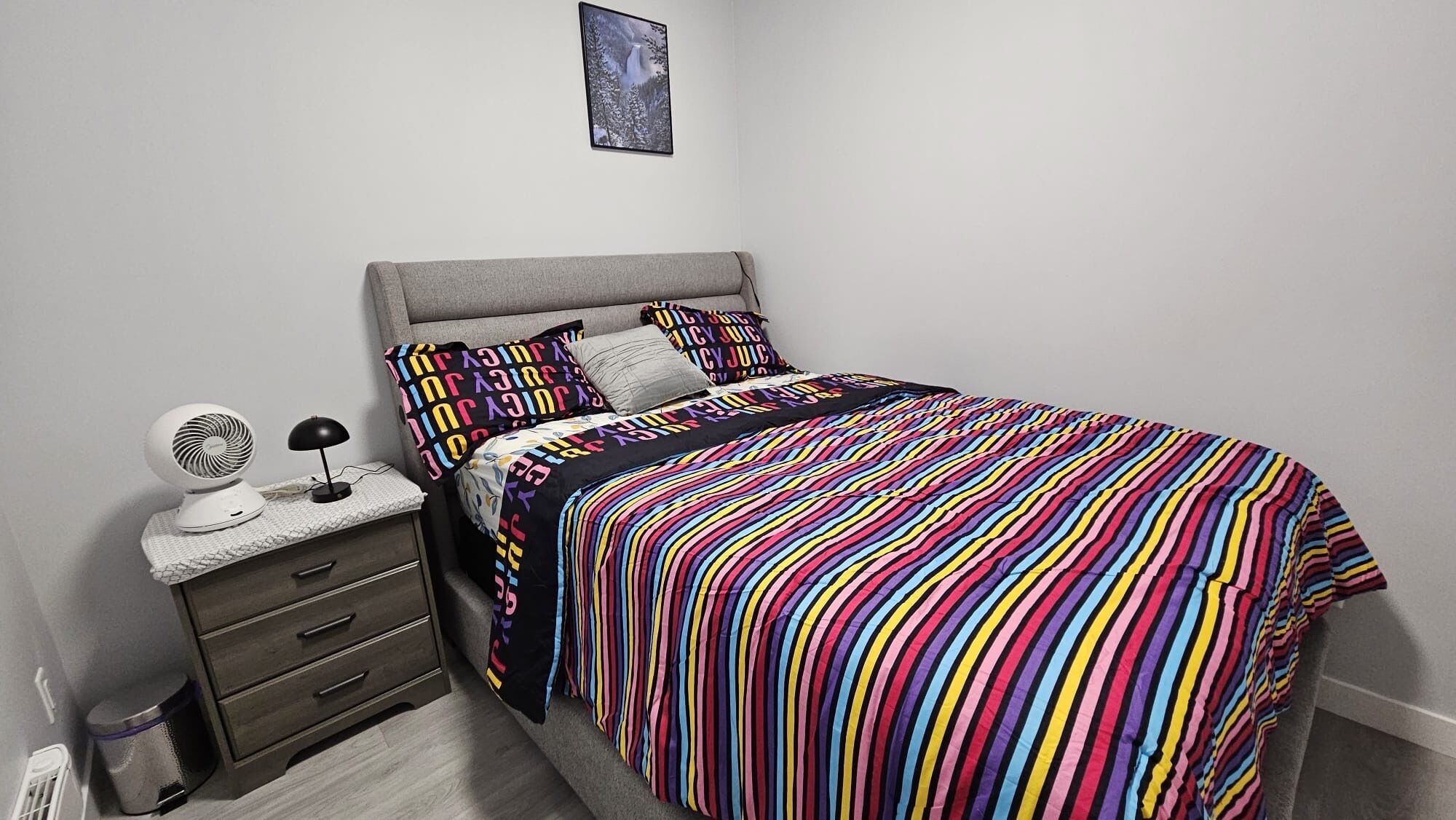 A bedroom with a bed, a bedside table with a fan, a lamp, and a trash bin.