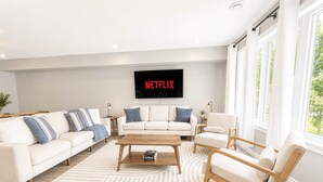 Un salon moderne avec un canapé blanc, une table basse en bois et un téléviseur fixé au mur qui affiche « NETFLIX ».