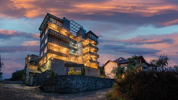 A multi-story building with balconies, a stone wall in front, and a dirt path leading to it.